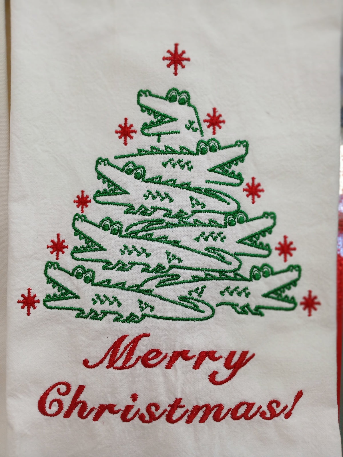 An embroidered Christmas towel featuring a tree made of green alligators, red snowflake accents, and the words 'Merry Christmas!' in red script on a white fabric background.